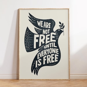 Peut inclure: Une affiche encadrée avec un motif de colombe bleu foncé et le texte blanc "WE ARE NOT FREE UNTIL EVERYONE IS FREE". La colombe tient une branche d'olivier. L'affiche est encadrée dans un cadre en bois.