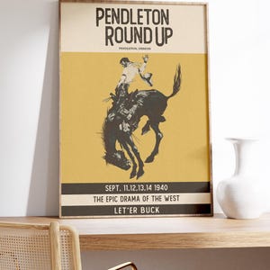 May include: Framed vintage poster for the Pendleton Round Up rodeo event. The poster features a black and white illustration of a cowboy riding a bucking bronco against a yellow background. Text includes event details.