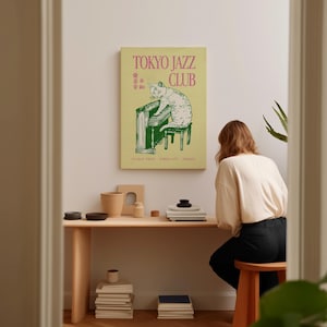May include: A framed poster with the text "TOKYO JAZZ CLUB" in pink and green, featuring a cat playing a piano. The poster is displayed on a white wall above a wooden desk with various decorative items.