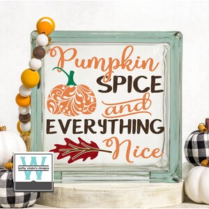 May include: A glass block with the text "Pumpkin Spice and Everything Nice" in brown and orange lettering. A stylized orange pumpkin with a swirl design is to the left of the text. A red and brown leaf with a stem is to the right of the text. The text "Kathy Winters Designs" is in a white square with a black border in the lower left corner.
