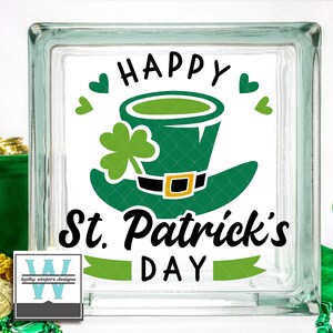 May include: A glass block with a white background and green shamrock design. The text "Happy St. Patrick's Day" is written in black lettering. A green leprechaun hat with a gold buckle is featured in the center of the design.