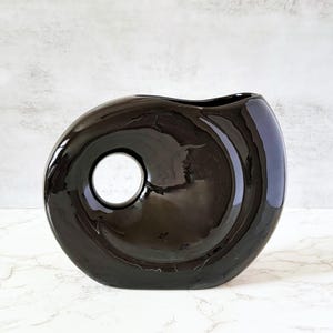 May include: A glossy, dark brown ceramic vase with a unique, rounded shape and a circular opening. The vase has a smooth, reflective surface and a small opening at the top. The design is modern and minimalist, suitable for home decor.