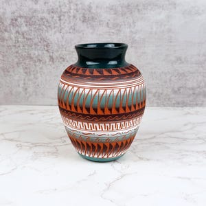 Könnte beinhalten: Eine Keramikvase mit dunkelgrünem Rand und bauchigem Körper, verziert mit horizontalen Bändern aus geometrischen Mustern in Terrakotta, Weiß und Türkis. Die Oberfläche zeigt wiederkehrende dreieckige und wellenförmige Motive.
