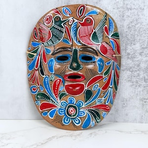 May include: An oval-shaped wooden mask featuring a hand-painted design of two birds at the top, a central face with a green nose and red lips, and a large blue flower at the bottom. The mask is decorated with vibrant red, green, and blue patterns.