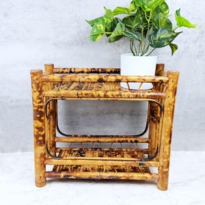 May include: A vintage bamboo plant stand with two shelves and a black metal frame. It features a white pot with a green and yellow leafy plant. The bamboo has a natural, warm tone with darker accents. The stand is a decorative piece for indoor plants.