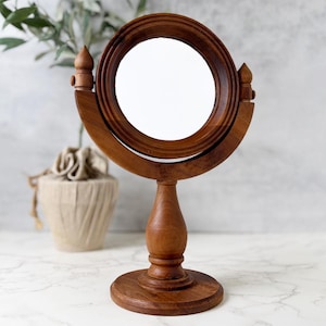 May include: A round tabletop mirror with a polished brown wooden frame and a matching wooden stand. The mirror is mounted on a swivel mechanism supported by a central pedestal base with a circular foot, featuring decorative finials on the sides.