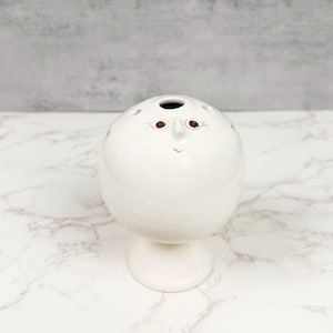 May include: A white ceramic bud vase with a whimsical face design. The vase has a round body with small holes and a single opening at the top. The face features brown eyes and a small, curved mouth. It sits on a pedestal base.