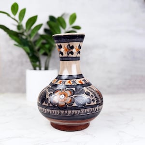 May include: A ceramic vase with a rounded body and a flared neck, featuring a beige base color decorated with hand-painted black and orange floral motifs, leaf patterns, and geometric bands encircling the neck and the widest part of the vessel.