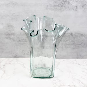 Può includere: Un vaso di vetro trasparente con la parte superiore a balze. Il vaso ha una base rettangolare e un corpo svasato. Il bordo a balze aggiunge un tocco decorativo. Il vaso è vuoto e poggia su una superficie di marmo.