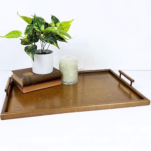 May include: A rectangular wooden serving tray with handles. The tray is a warm brown color and has a smooth surface. A small white potted plant, two books, and a glass jar with a lid are on the tray.