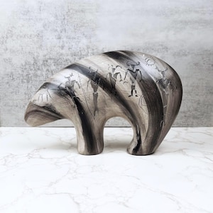 Puede incluir: Figura de oso de cerámica en tonos grises y negros. El oso tiene un cuerpo redondeado y está decorado con figuras y patrones estilizados. La obra de arte parece representar antiguas pinturas rupestres. La figura se exhibe sobre una superficie de mármol blanco.