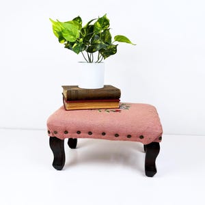 May include: A small, antique footstool with a pink and white checkered fabric top. The top is adorned with embroidered floral details and dark brown nailhead trim. Dark wooden legs support the stool. A stack of old books and a potted plant sit on top.
