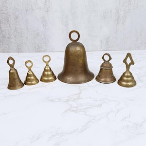 May include: A collection of six antique brass bells of varying sizes and shapes. The bells range from small, conical designs to a large, rounded bell with a loop handle. The brass has a warm, aged patina, and the bells are arranged on a white marble surface.
