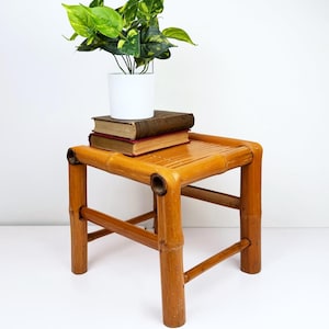 May include: A small, square bamboo side table with a natural finish. The table has a slatted top and rounded legs. A white ceramic pot with a green and yellow plant sits on top of two books. The table is approximately 15 inches tall.