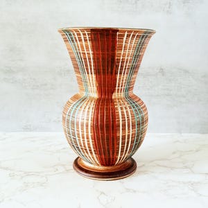 Puede incluir: Un jarrón decorativo de cerámica con cuerpo bulboso y parte superior acampanada. El jarrón presenta rayas verticales en tonos marrón, naranja, blanco y turquesa. Tiene una base redonda y está sobre una superficie de mármol.