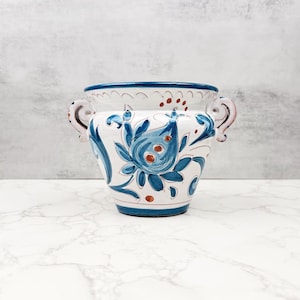 Puede incluir: Un jarrón de cerámica blanco con diseños florales azules y dos asas. El jarrón tiene una abertura ancha y un borde azul. El diseño floral incluye hojas azules y detalles en naranja. El jarrón es probablemente para fines decorativos.