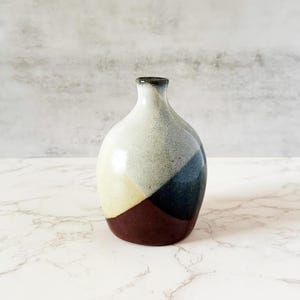May include: A small ceramic vase with a rounded body and a narrow neck. The vase is divided into colour blocks of cream, yellow, blue, and brown. The vase is likely handmade and has a glossy finish. The vase is a decorative item.