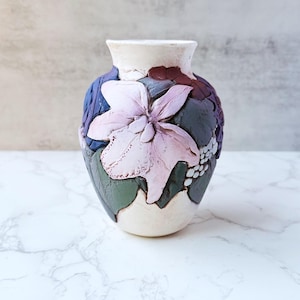 May include: A cream-colored ceramic vase with a floral design. The vase features a large, raised pink flower and other raised floral elements in shades of blue, green, and purple. The vase has a wide opening and a textured surface.