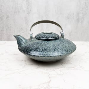 Könnte beinhalten: Eine blau-graue Keramik-Teekanne mit einem silbernen Metallgriff und Deckel. Die Teekanne hat einen abgerundeten Körper und einen Ausguss. Der Deckel ist flach und kreisförmig. Die Teekanne steht auf einer weißen Marmoroberfläche.