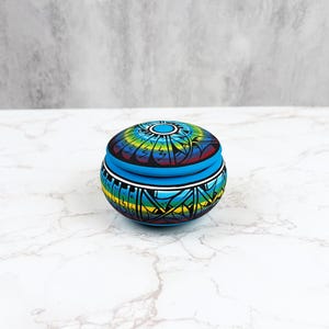 Puede incluir: Pequeña caja redonda para baratijas con tapa. La tapa y los lados están decorados con un colorido patrón geométrico en tonos azules, verdes, amarillos y rojos. La caja está sobre una superficie de mármol blanco.