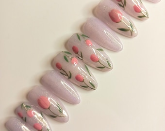 May include: A set of lavender-colored, almond-shaped artificial nails. Each nail features a hand-painted design of pink tulips with green leaves. The nails have a glittery finish, adding a touch of sparkle to the floral design.