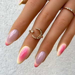 Peut inclure: Gros plan sur une main ornée de plusieurs bagues dorées et de longs ongles ovales. Les ongles présentent un dégradé de rose à jaune. Une bague arbore un motif de croissant de lune et d'étoile.