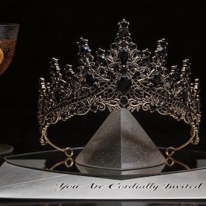 May include: A black and gold crown with dark gemstones. The crown is displayed on a mirrored surface with the text "You Are Cordially Invited". The crown has a vintage, regal aesthetic, suitable for special occasions.