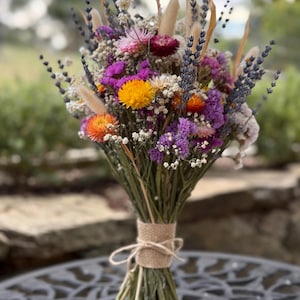 May include: A colourful bouquet of dried flowers, with orange, yellow, purple, and white blooms. The stems are bound with twine. The arrangement is set against a blurred outdoor background.
