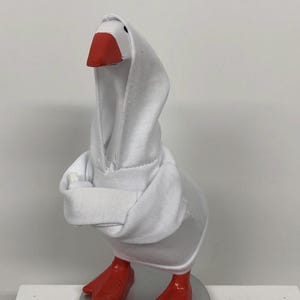 May include: A white goose figurine wearing a white hooded sweatshirt. The goose has a red beak and red feet. The figurine is standing on a silver-colored circular base.