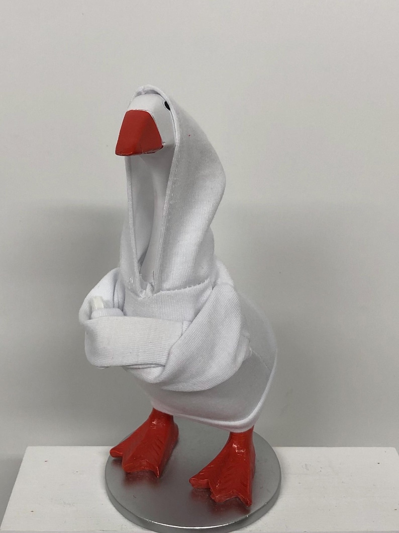 May include: A white goose figurine wearing a white hooded sweatshirt. The goose has a red beak and red feet. The figurine is standing on a silver-colored circular base.