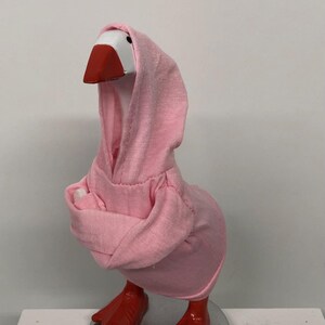 May include: A whimsical goose figurine wearing a pink hoodie. The goose has a white head, a red beak, and red feet. The figurine is standing on a silver-colored base. The pink hoodie covers the goose's body and head, creating a playful aesthetic.