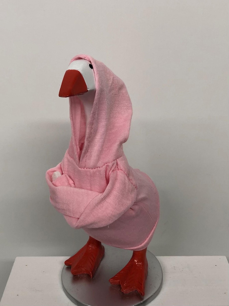 May include: A whimsical goose figurine wearing a pink hoodie. The goose has a white head, a red beak, and red feet. The figurine is standing on a silver-colored base. The pink hoodie covers the goose's body and head, creating a playful aesthetic.