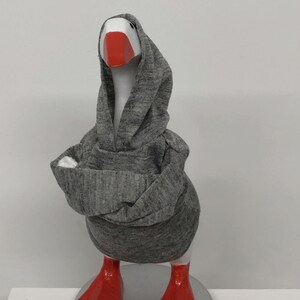 May include: A whimsical goose figurine wearing a gray hoodie. The goose has a white head, a bright red beak, and matching red feet. The figurine is standing on a silver-colored base, creating a fun and quirky decorative item.