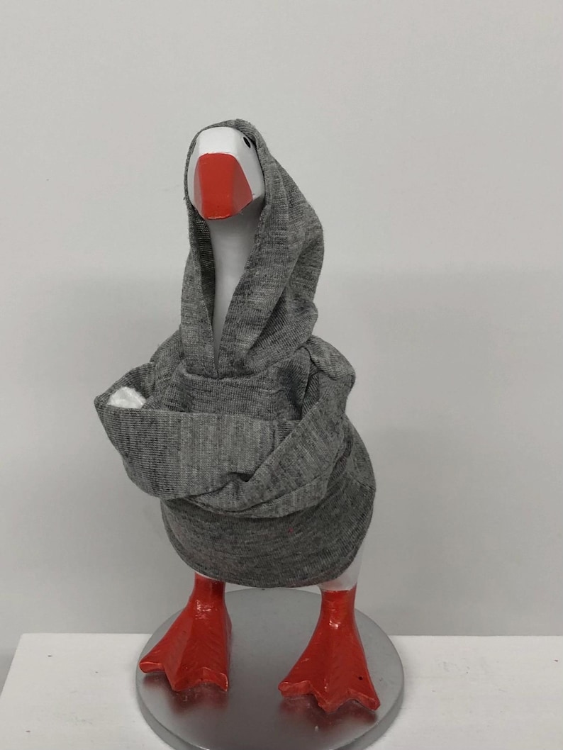 May include: A whimsical goose figurine wearing a gray hoodie. The goose has a white head, a bright red beak, and matching red feet. The figurine is standing on a silver-colored base, creating a fun and quirky decorative item.