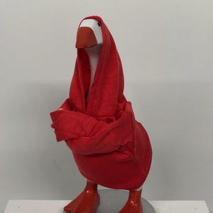 May include: A whimsical goose figurine with a white head, orange beak, and red webbed feet. The goose is draped in a red hooded garment, giving it a unique and playful appearance. The figurine is mounted on a silver-colored circular base.