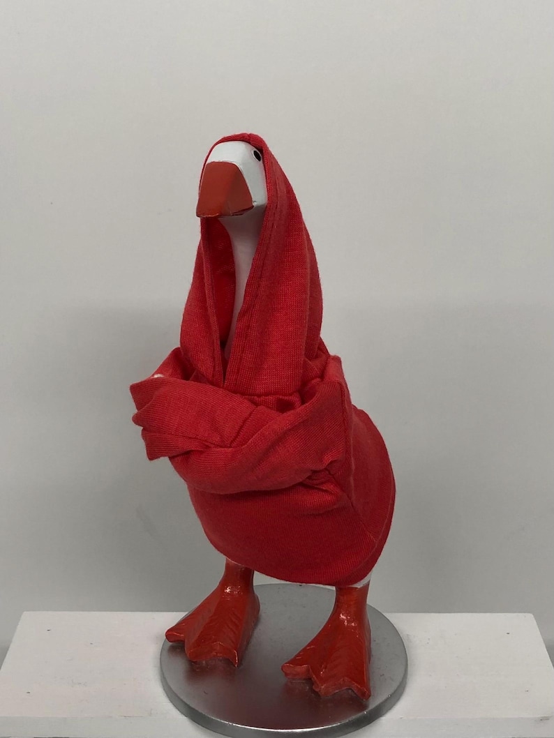 May include: A whimsical goose figurine with a white head, orange beak, and red webbed feet. The goose is draped in a red hooded garment, giving it a unique and playful appearance. The figurine is mounted on a silver-colored circular base.