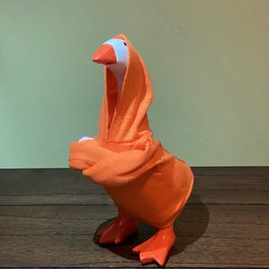 May include: A whimsical goose figurine wearing an orange hooded robe. The goose has a white head, an orange beak and feet. The robe is draped over the goose's body, creating a playful and unique appearance. The background is a solid green.