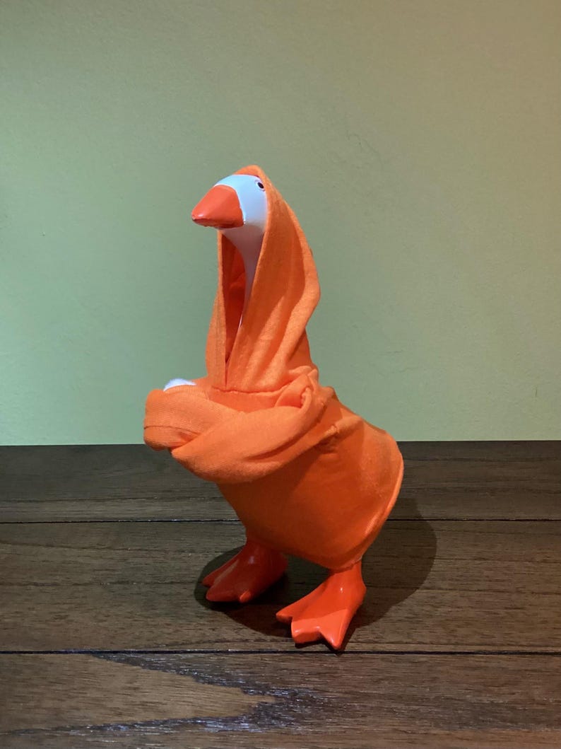 May include: A whimsical goose figurine wearing an orange hooded robe. The goose has a white head, an orange beak and feet. The robe is draped over the goose's body, creating a playful and unique appearance. The background is a solid green.
