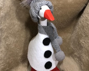 May include: A white goose figurine dressed as a snowman with a gray knitted hat, scarf, and pom-poms. The goose has a red beak and red feet.
