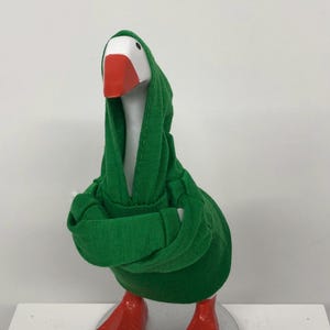 May include: A whimsical goose figurine with a white head, orange beak and feet, and a green hooded sweatshirt. The goose is standing on a silver base, with its arms crossed. The figurine is a fun and unique decorative item.