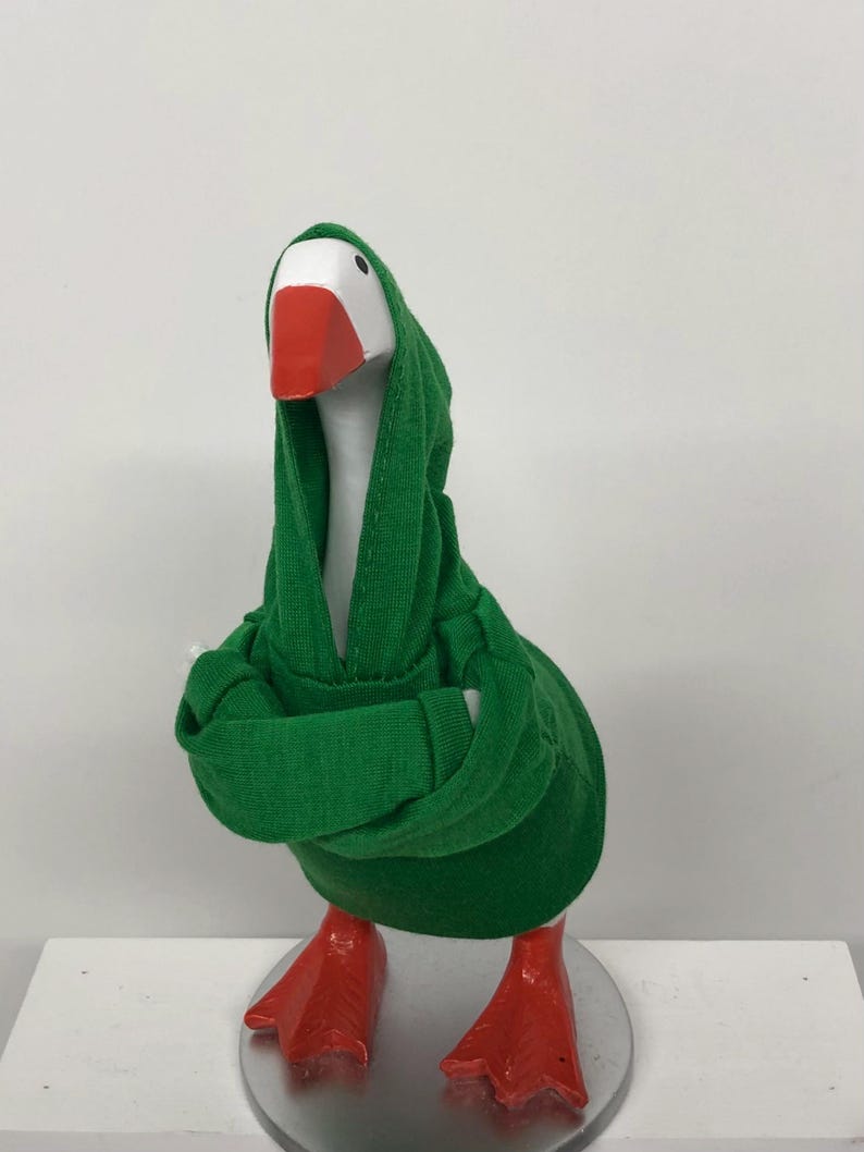 May include: A whimsical goose figurine with a white head, orange beak and feet, and a green hooded sweatshirt. The goose is standing on a silver base, with its arms crossed. The figurine is a fun and unique decorative item.