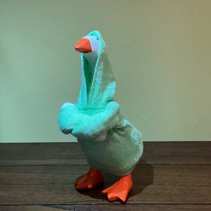 May include: A plush toy goose wearing a light green hooded robe. The goose has a white head, an orange beak, and orange feet. The background is a solid light green color, and the goose is standing on a wooden surface.
