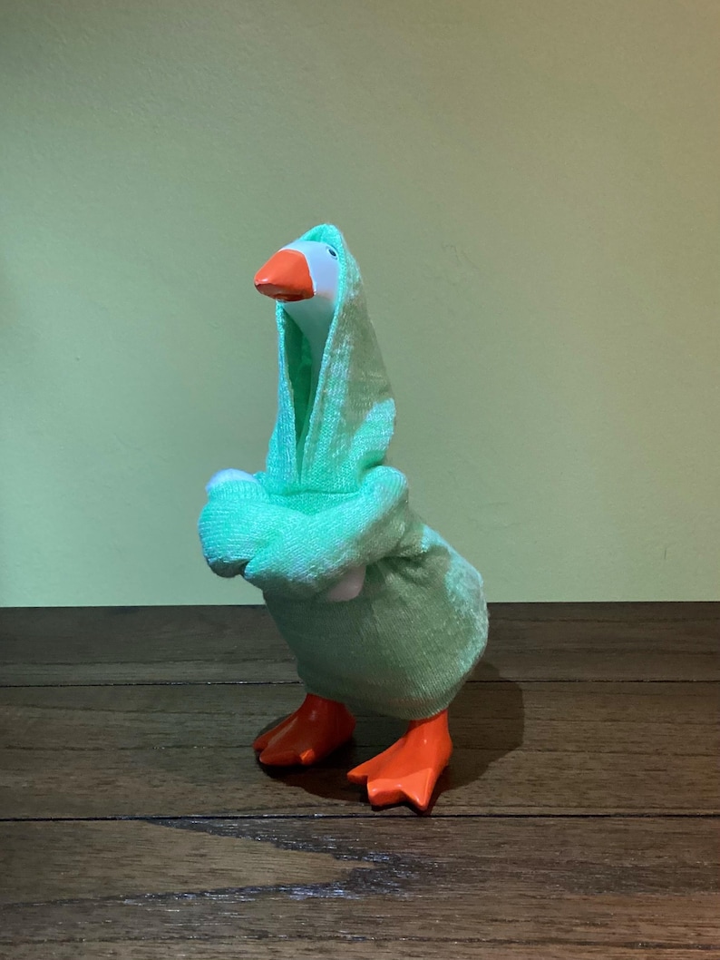 May include: A plush toy goose wearing a light green hooded robe. The goose has a white head, an orange beak, and orange feet. The background is a solid light green color, and the goose is standing on a wooden surface.