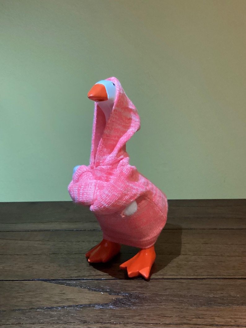 May include: A whimsical goose figurine with an orange beak and feet, wearing a pink hooded sock. The goose has a white head and is standing on a wooden surface. The background is a solid green color.