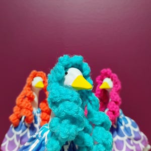 Può includere: Tre fantasiose statuette di oche con teste bianche, becchi gialli e capelli colorati in blu, arancione e rosa. Indossano abiti a fantasia blu e bianchi con dettagli viola, su uno sfondo viola scuro.