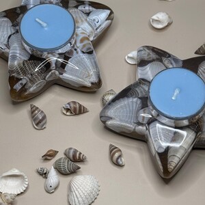 May include: Two star-shaped candle holders made of seashells, each holding a light blue candle. Various seashells are scattered around the candle holders. The candle holders are filled with a variety of seashells in shades of white, brown, and tan.