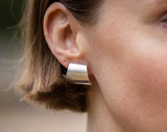 May include: A close-up of a silver-colored, square-shaped earring. The earring has a slightly angled, geometric design. The earring is worn on the ear of a person with short, brown hair.