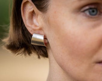 May include: A close-up of a silver earring with a wavy, textured design. The earring is a square shape that curves around the earlobe. The earring is made of a shiny metal and reflects light.