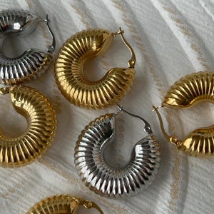 May include: Close-up of several gold and silver hoop earrings. The earrings have a ribbed, textured design and a curved shape. The earrings are displayed on a white, textured surface.
