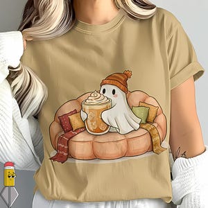 Peut inclure: T-shirt beige avec un dessin d'un fantôme de dessin animé portant un bonnet en tricot, se relaxant sur un canapé en forme de citrouille avec des coussins et une écharpe. Le fantôme tient un verre de café glacé à la crème fouettée.